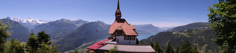 Panoramablick über das Dach einer Kirche auf bewaldete Hügel und schneebedeckte Berge.