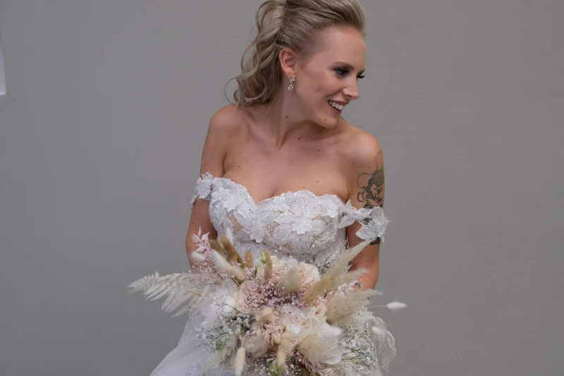 Eine lächelnde Braut mit Blumenstrauss vor einem ruhigen grauen Hintergrund.