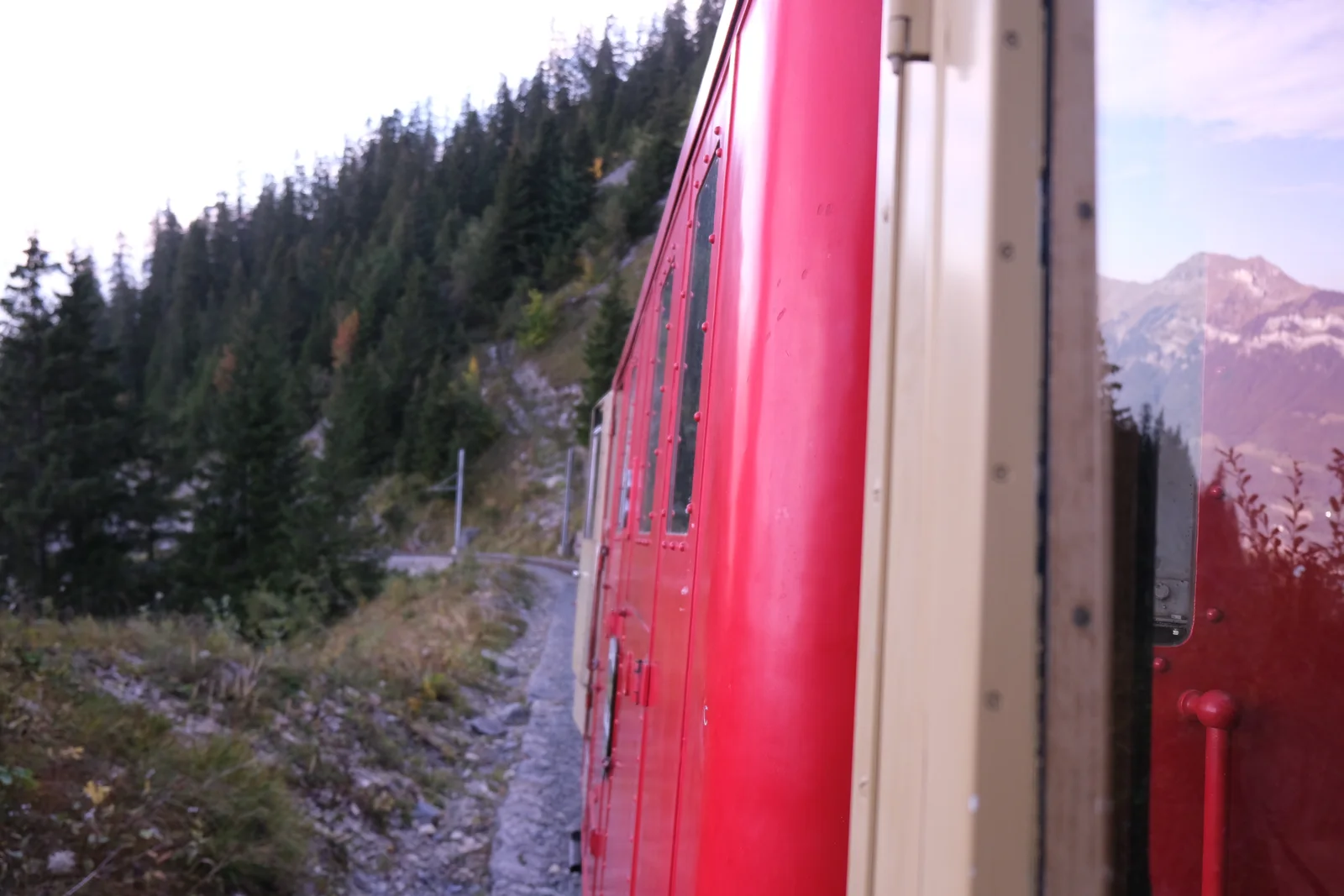 Blick entlang eines roten Zuges auf Gleise, Wald und Berge.